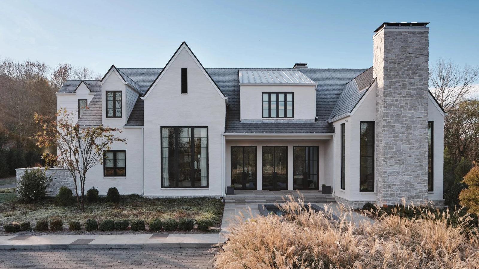 A modern two-story house exterior with white stucco walls, dark-framed windows, a gray metal roof, and a tall light stone chimney. The property has a paved driveway, landscaping, and dry ornamental grasses in the foreground under a clear blue sky.