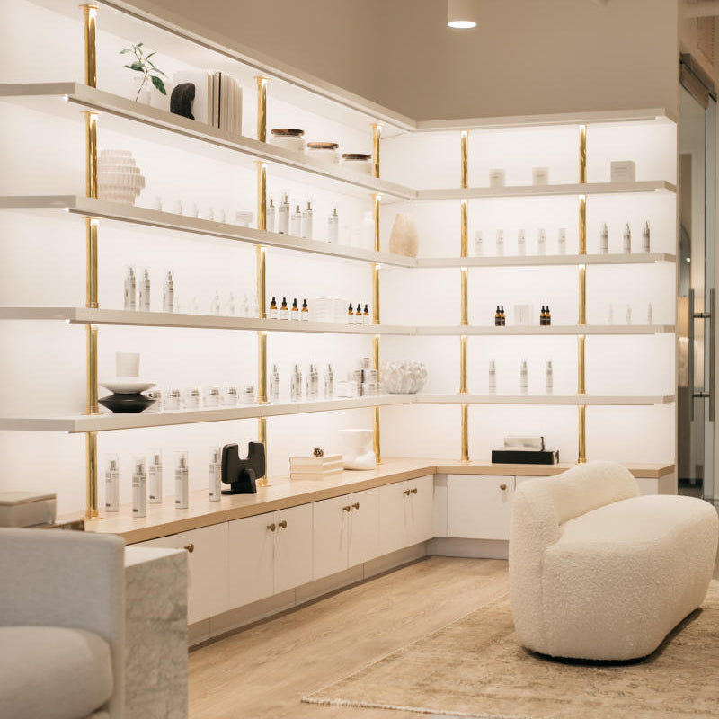 A modern, bright retail interior with white built-in shelving and gold trim displaying many skincare or beauty products. A cream boucle lounge chair sits on a light rug, with part of a sofa and a marble coffee table visible in the foreground.