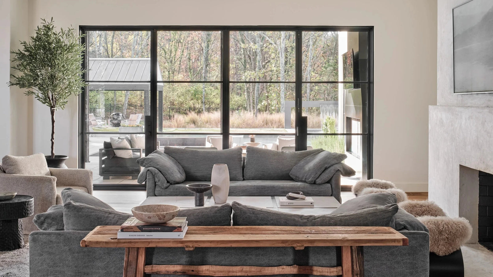 A modern living room featuring a gray fabric sofa set, a rustic wooden coffee table, a white fireplace, and large black-framed windows looking out onto trees.