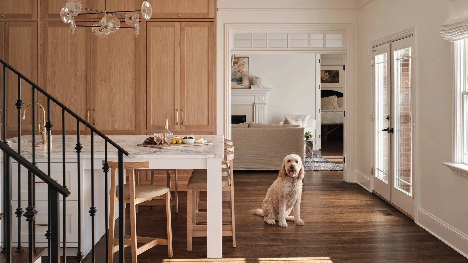 A golden retriever sits on a dark wood floor in a bright, modern kitchen and dining area with light oak cabinets, a white marble island, and a black metal staircase railing.