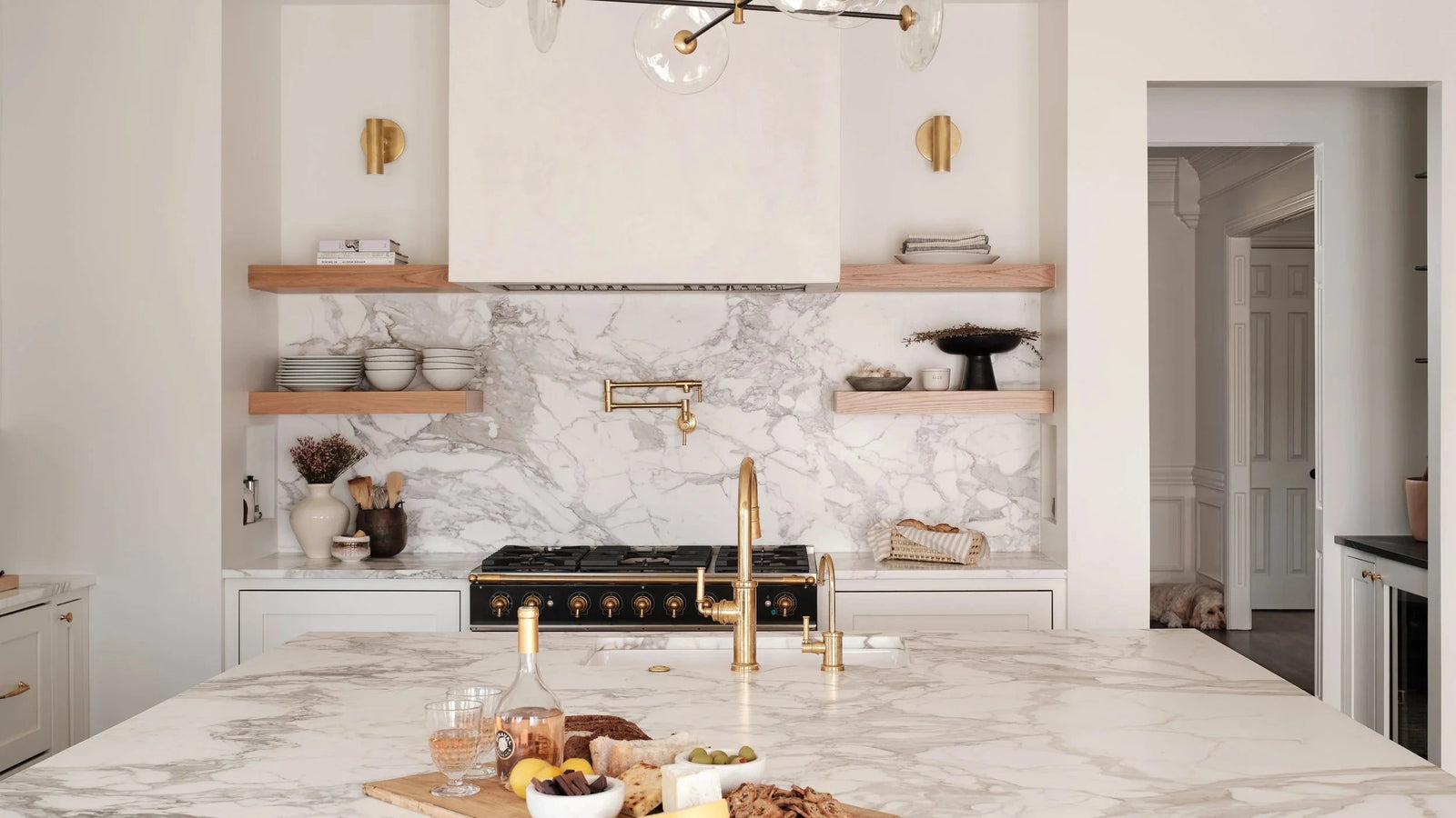 A modern kitchen island with a white and gray marble surface, matching marble backsplash, gold faucet, and a black and gold chandelier with clear glass globes above.
