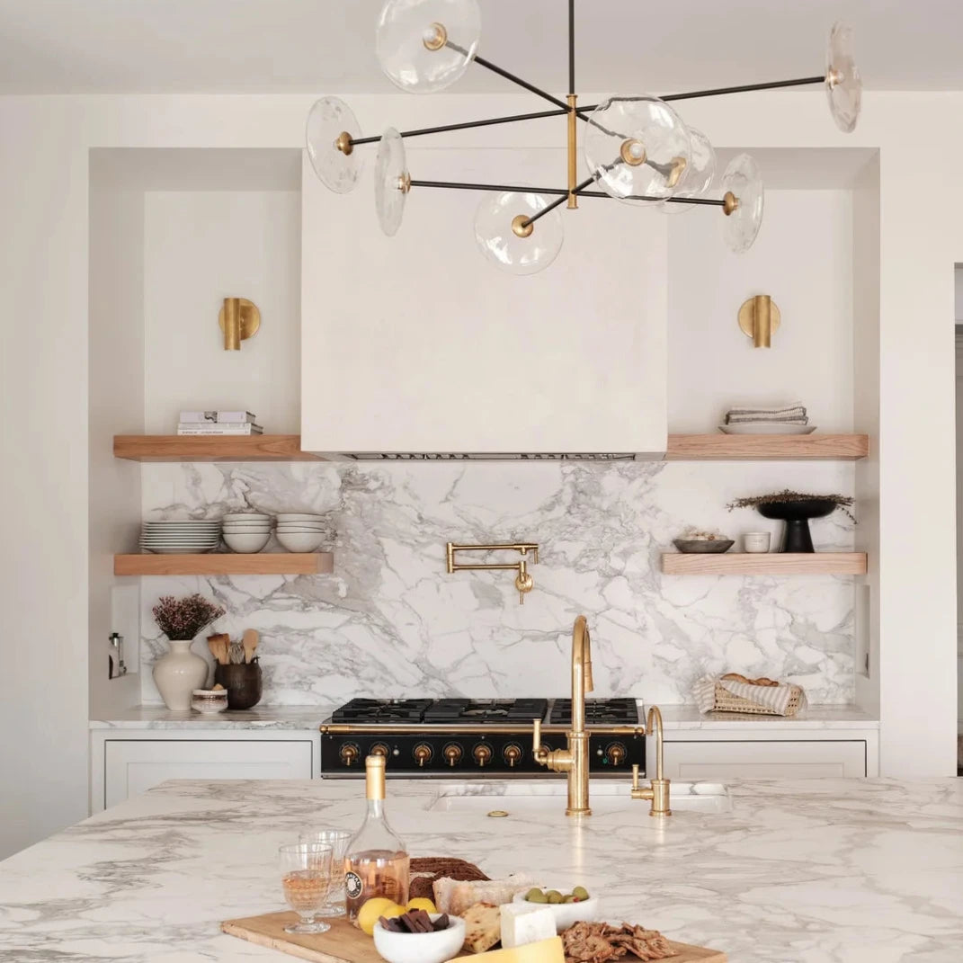 A modern kitchen island with a white and gray marble surface, matching marble backsplash, gold faucet, and a black and gold multi-arm chandelier with clear glass globes hanging above.