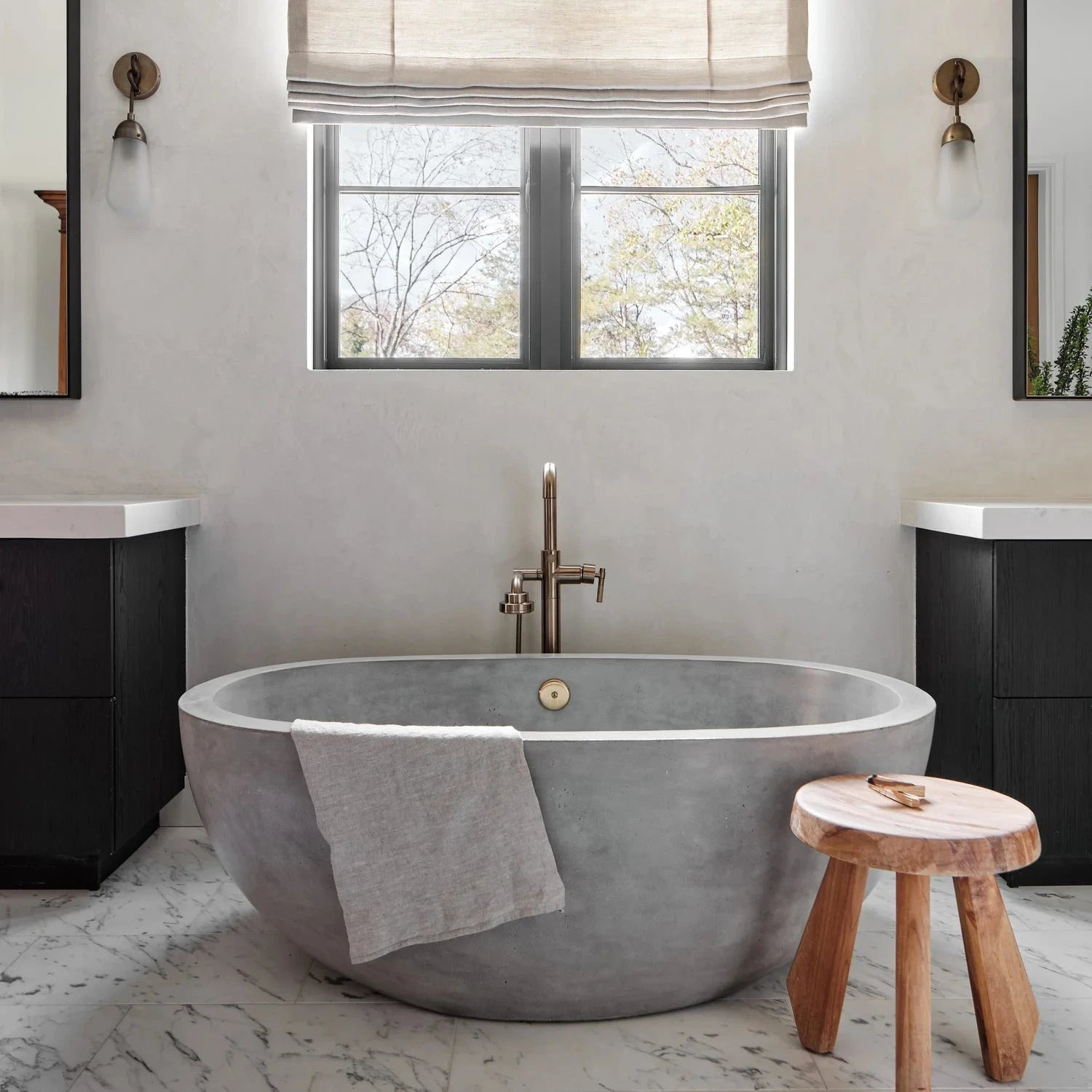 A round concrete freestanding bathtub in a modern bathroom setting with a window, beige roman shade, black vanity cabinets, and a small wooden stool.