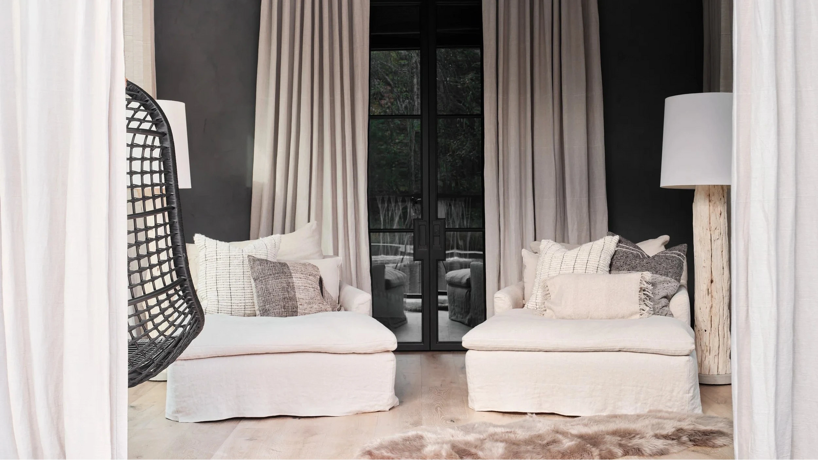 A styled interior scene showing two off-white slipcovered armchairs with pillows, draped beige curtains, a dark charcoal wall, a black hanging egg chair, and floor lamps.