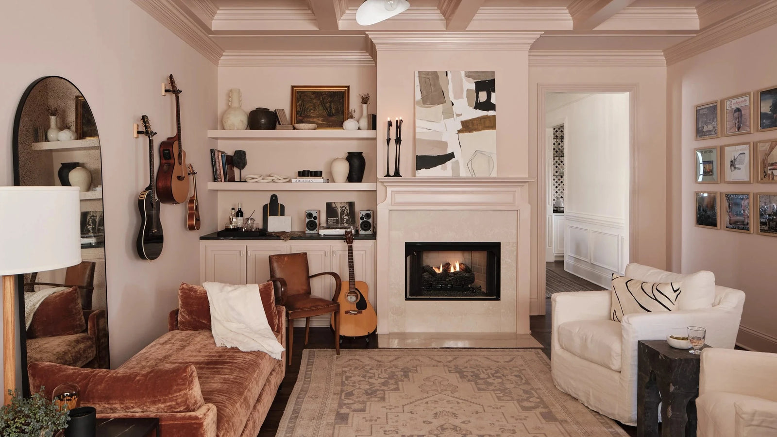 A beautifully styled living room featuring a coffered ceiling, a built-in fireplace with shelves, a rust-brown velvet sofa, a white armchair, and several acoustic guitars hanging on the wall.