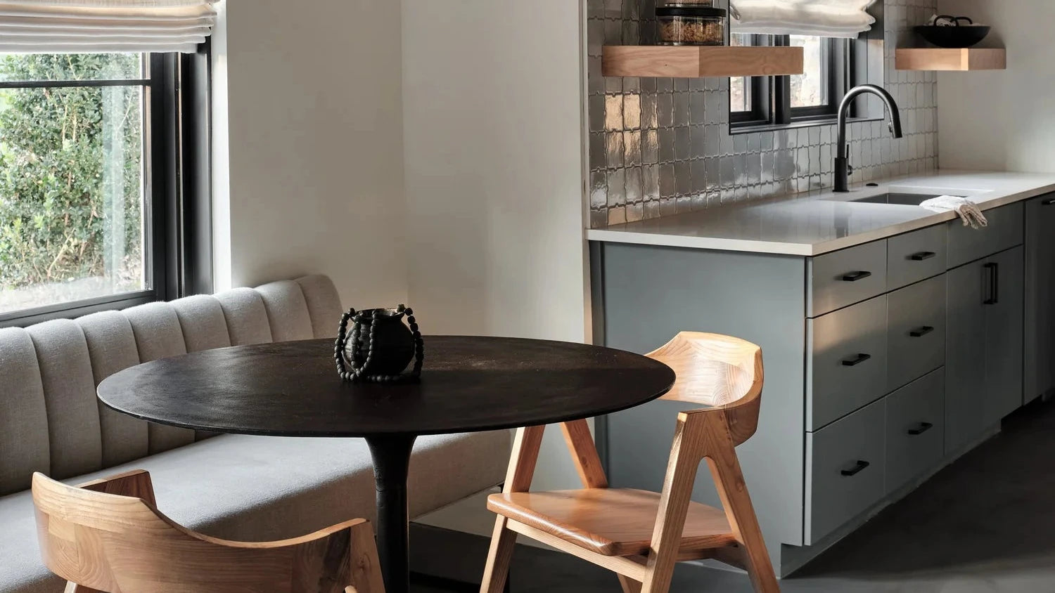 A modern dining nook with a round black table, two light wood chairs, and a gray upholstered banquette under a window. In the background is a contemporary kitchen with gray cabinets, a tiled backsplash, and floating wooden shelves.