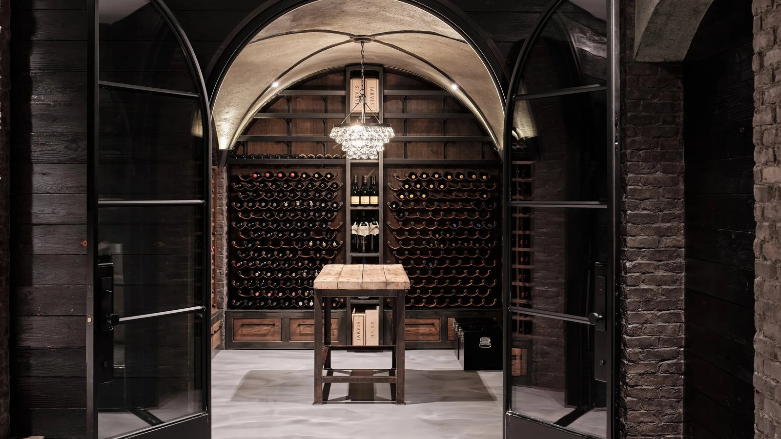 A luxury wine cellar interior with a central wooden work table, a hanging crystal chandelier, and extensive wine bottle racks under a stone barrel vault, framed by open black double doors.