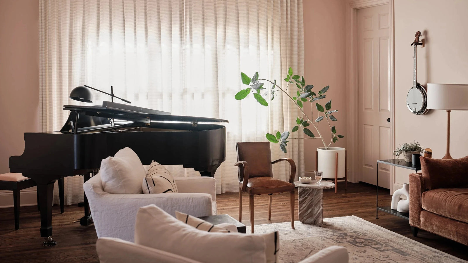A styled living room interior with blush pink walls, sheer white curtains, a black grand piano, cream upholstered seating, a brown accent chair, a side table with a marble top, and a potted green plant.