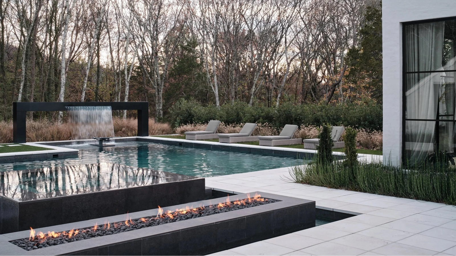 Modern outdoor poolside patio with a rectangular fire pit in the foreground, a black-framed waterfall feature, lounge chairs, bare trees, and part of a white house with large windows.