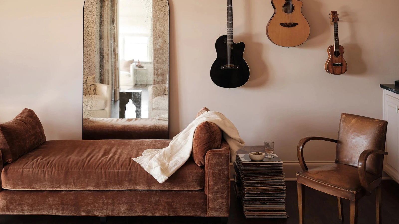 A styled interior scene with a brown velvet chaise lounge, a tall arched mirror, wall-mounted acoustic and ukulele guitars, a leather armchair, and a stack of records beside the chair.