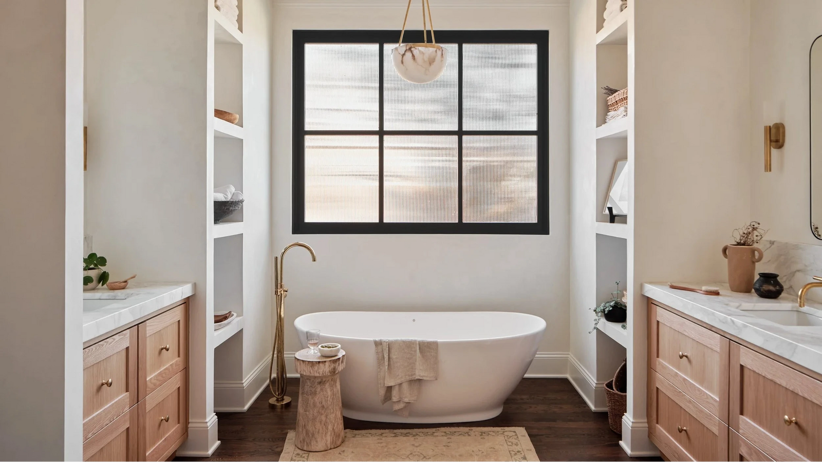 A luxury bathroom interior featuring a freestanding oval bathtub centered beneath a black-framed window. The room has warm wood cabinetry, white marble countertops, built-in shelving, a hanging pendant light, and dark wood flooring.