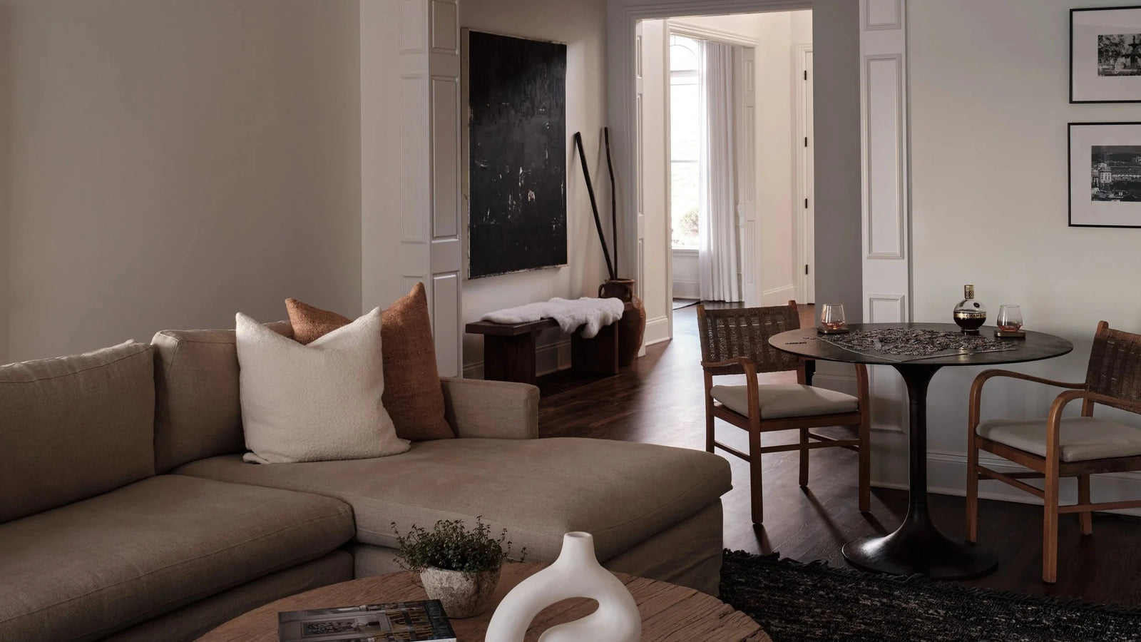 A modern living room featuring a taupe sectional sofa with white and brown accent pillows, a round wooden coffee table with a white sculptural vase, and a dining area in the background.