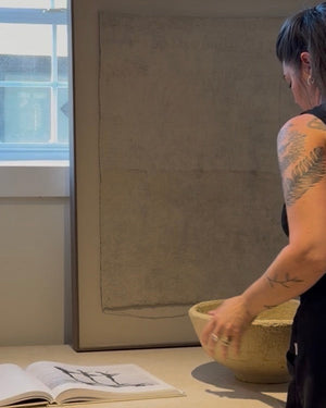 A person with tattoos stands beside a large framed abstract artwork in a bright room, reaching toward a woven bowl on a table. An open book and wooden chair are visible in the foreground.