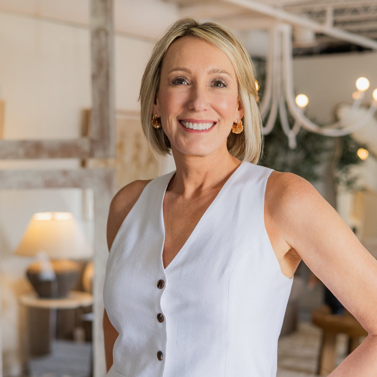 A smiling blonde woman wearing a white sleeveless button-front vest and light blue jeans, standing in a softly lit home decor or furniture showroom.