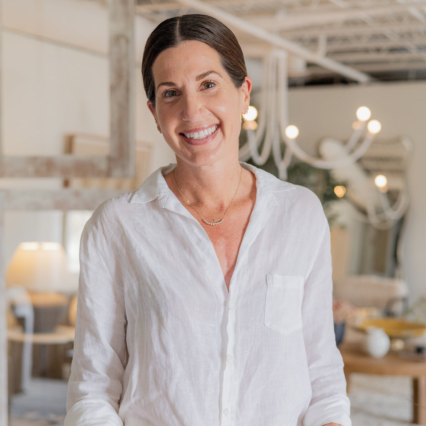 A smiling woman with dark hair pulled back wears a loose white long-sleeve button-up shirt and white pants, standing in a bright, modern home decor showroom with furniture and string lights blurred in the background.