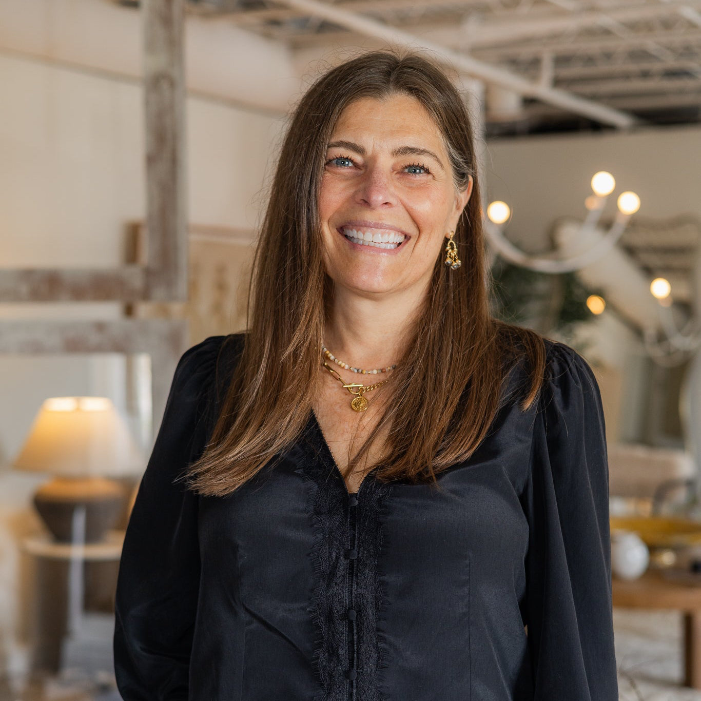 A smiling woman with long brown hair stands indoors wearing a black long-sleeve satin blouse with gathered cuffs and white pants. The background shows a softly blurred home decor or furniture interior with lamps and decor.