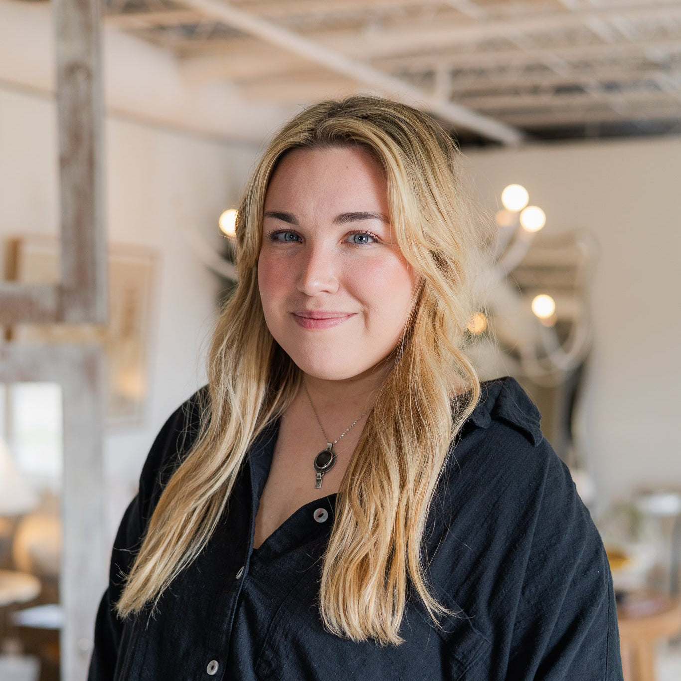 A smiling blonde woman wearing a loose black button-up shirt stands in a bright, softly blurred indoor room with exposed ceiling beams and hanging lights.