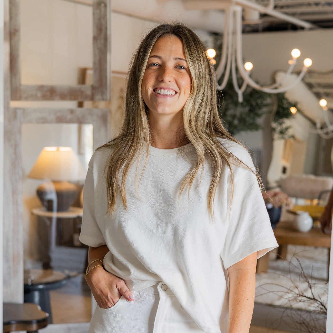 A smiling woman with blonde hair stands in a bright, rustic home decor showroom wearing a matching off-white short-sleeve top and high-waisted trousers.