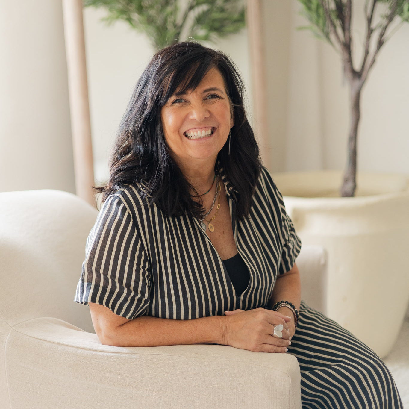 A smiling woman with dark hair sits on a cream upholstered chair in a bright indoor setting, wearing a black and white vertically striped short-sleeve top and matching striped pants.