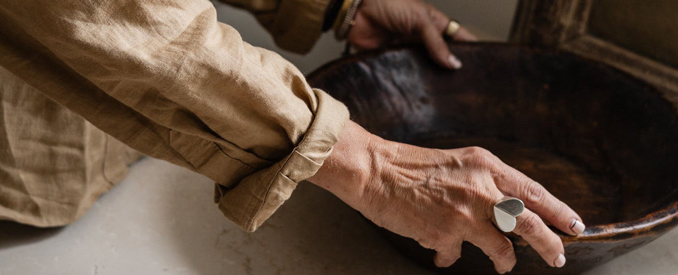 A person in a beige linen shirt holds a large dark brown wooden bowl on a light stone countertop in a warm, neutral-toned room.