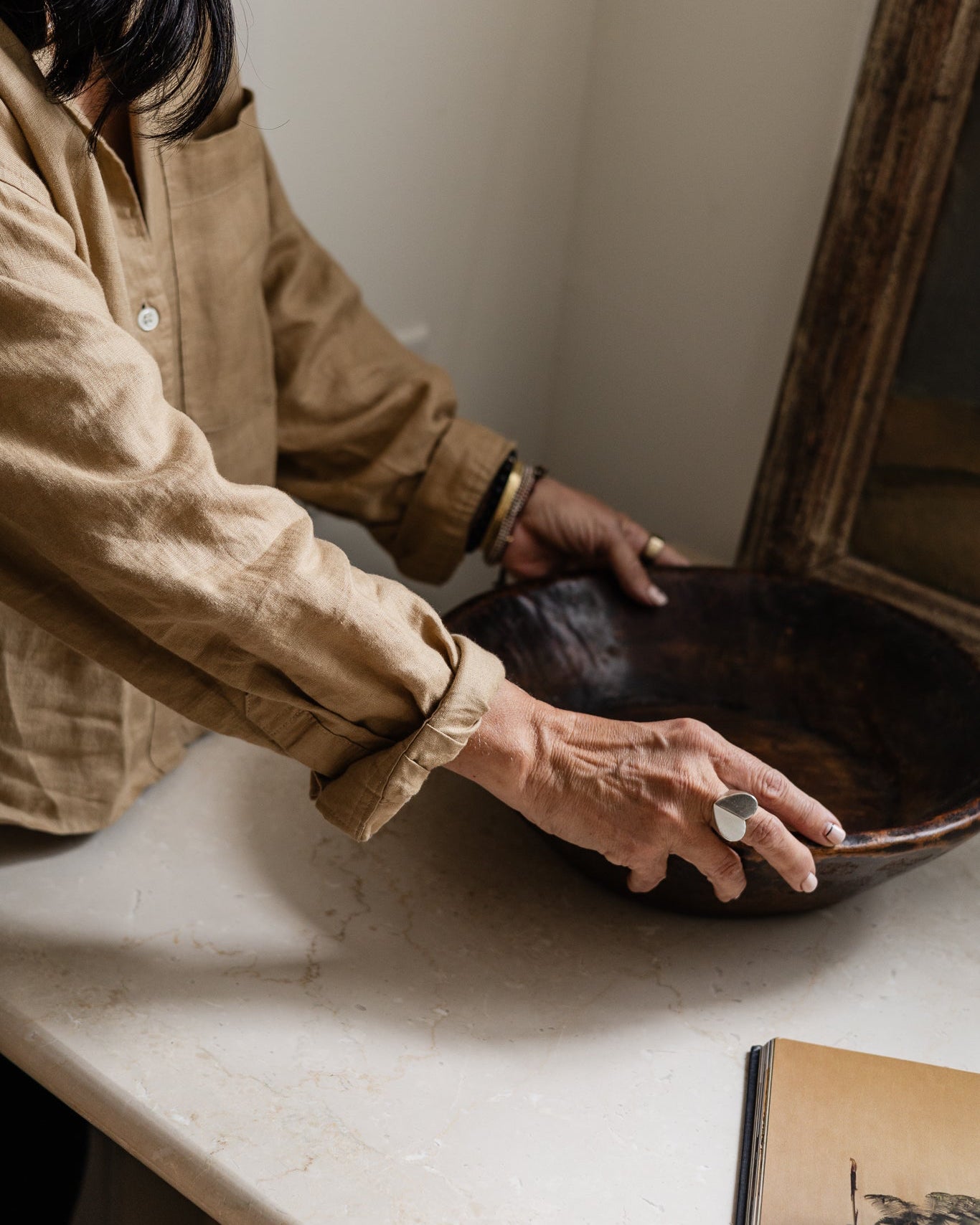 A person in a beige linen shirt holds a large dark brown wooden bowl on a light stone countertop in a warm, minimalist interior setting.