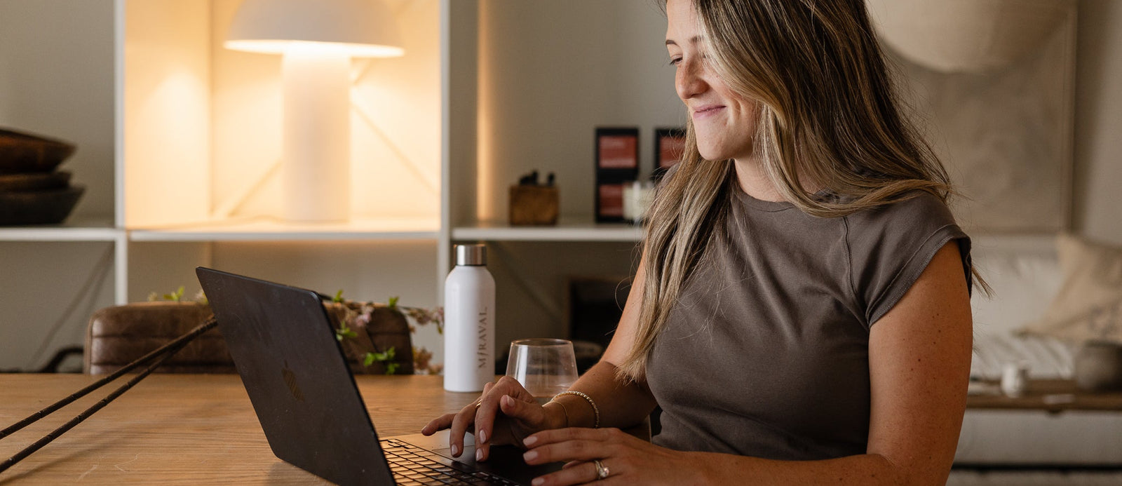 A woman with long light brown hair sits at a wooden table in a modern home office, smiling as she uses a black laptop. She is wearing a taupe short-sleeve top and beige pants.