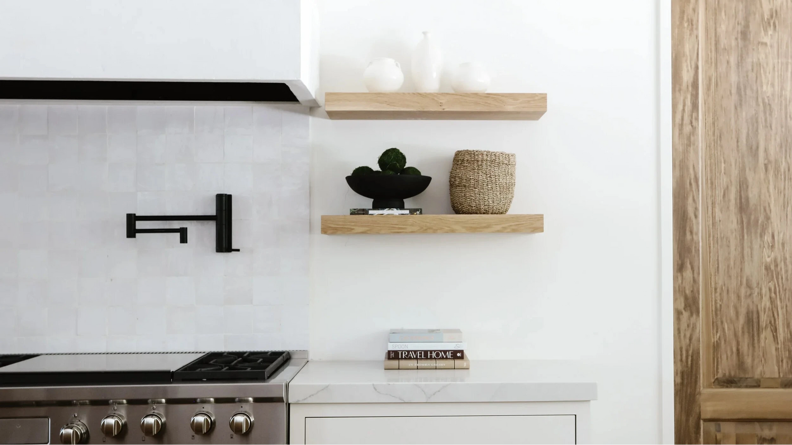 A modern kitchen interior with two light wood floating shelves mounted on a white wall. The shelves hold white ceramic vases, a small black decorative object, and a woven basket.