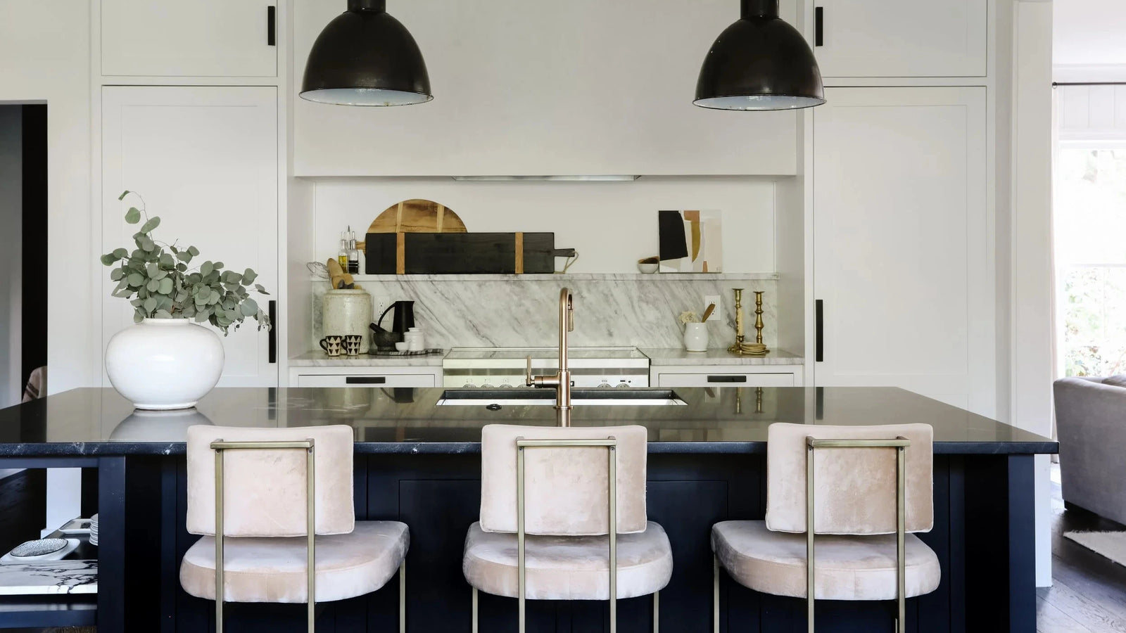 A modern kitchen island with a dark wood base and black countertop, featuring three blush pink velvet bar stools with gold metal legs. The background shows white cabinetry, a marble backsplash, and two black dome pendant lights hanging above.