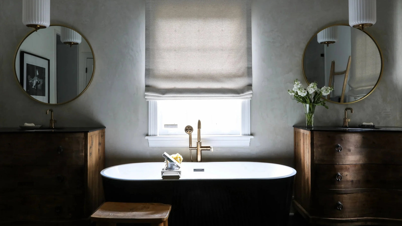 A classic freestanding white bathtub with a gold faucet in a rustic bathroom setting, flanked by dark wood dressers, round mirrors, and a window with a beige roller blind.