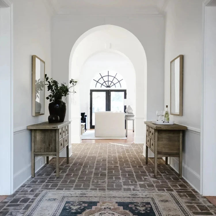 A long hallway with white walls, arched doorways, a geometric brass chandelier, and a large patterned area rug in blue, beige, and brown tones. Two wooden console tables with mirrors and a black vase with flowers sit on either side.