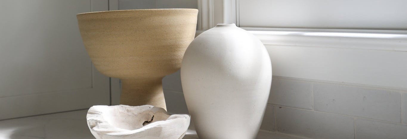 A collection of minimalist ceramic and wood home decor items, including a light wood bowl, a white ceramic vase, and a small white dish, arranged on a white windowsill next to a bright window.