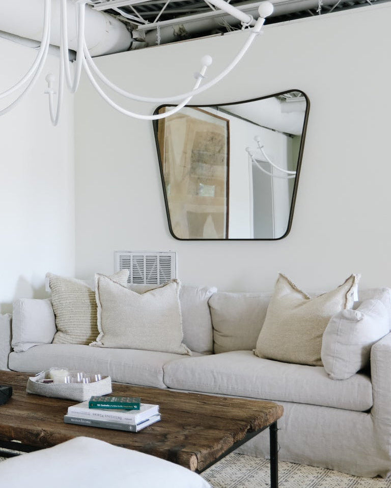 A modern living room featuring a light beige fabric sofa with neutral throw pillows, a rustic dark wood coffee table, and a large, uniquely shaped black-framed mirror on a white wall.