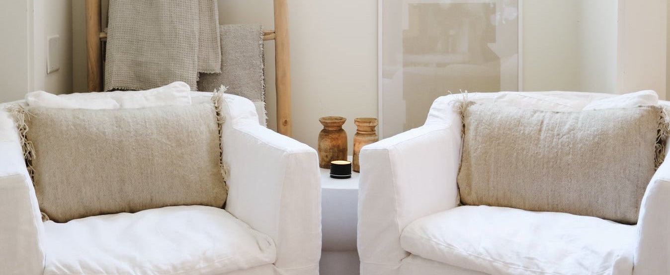A bright, minimalist room featuring two cream-colored slipcovered armchairs facing each other, with neutral-toned pillows, a wooden ladder shelf with folded textiles, and a window in the background.