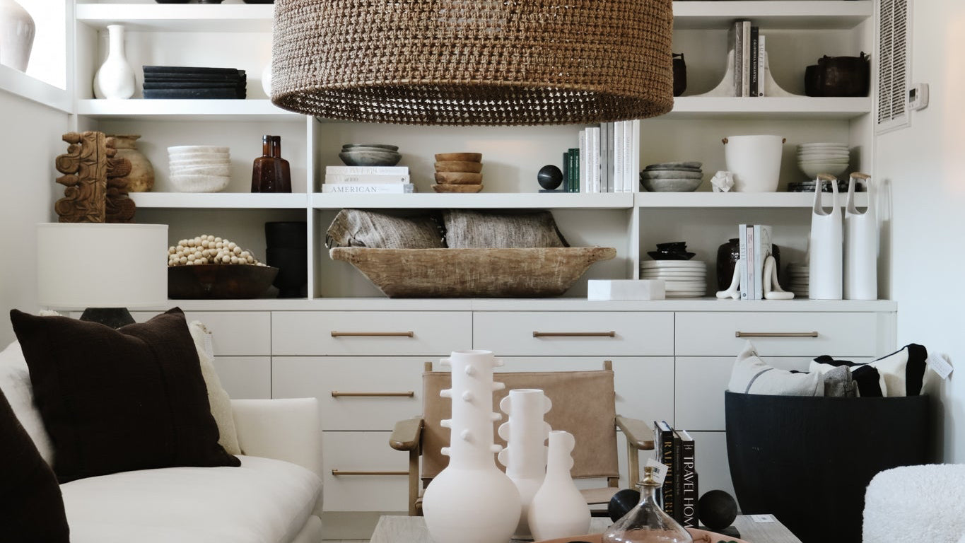 A large woven wicker pendant light hangs in a bright, modern living room with a white sofa, a low wooden coffee table, and built-in white shelving in the background.