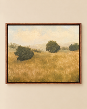 A framed landscape painting of a golden grassy field with three dark green trees under a pale sky, displayed on a beige wall.