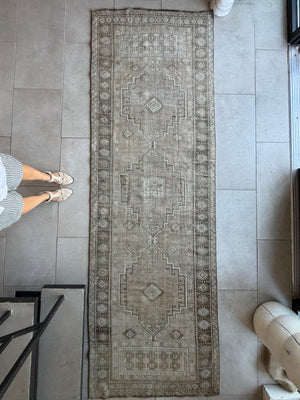 A long, narrow vintage-style runner rug with a distressed geometric pattern in muted beige and gray tones, laid out on a light gray tiled floor. A person's legs and shoes are visible on the left side of the frame.