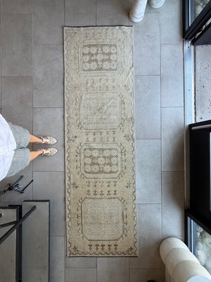 A long rectangular vintage-style runner rug with a distressed beige and gray geometric pattern, laid out on a gray tiled floor. A person's legs and feet are visible on the left side of the frame.
