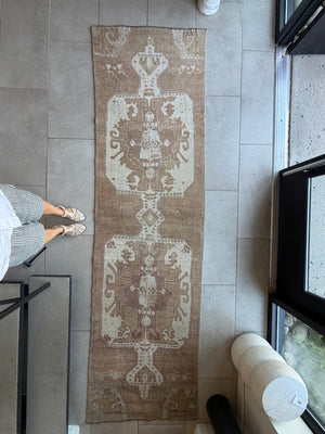 A long rectangular area rug with an ornate distressed medallion pattern in beige, brown, and cream, laid out on a gray tiled floor. A person's legs and feet are visible on the left side of the frame, and a window and white cylindrical object are visible on the right.