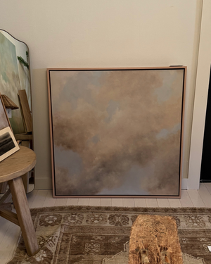 A large framed abstract painting with soft beige, taupe, and gray brushstrokes leans against a cream wall in a styled room with a rug, stool, and mirror visible.