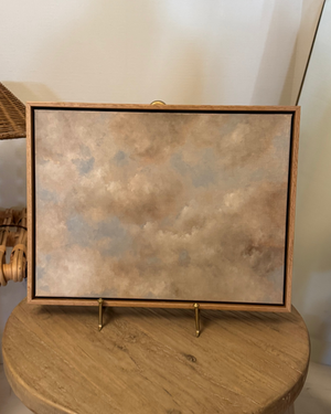 A framed art print featuring a textured, cloudy abstract design in earthy beige, brown, and muted blue-gray tones, displayed on a wooden stool.