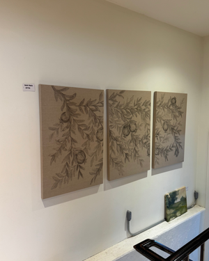 Three vertical canvas panels with botanical illustrations of branches and fruit hang on a beige wall in an indoor gallery or showroom. A small label is visible near the left panel, and part of a staircase railing and a small landscape painting appear in the foreground and lower right.
