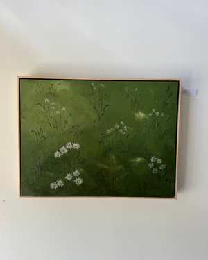 A framed painting depicting a green meadow with small white wildflowers, hung on a plain white wall.