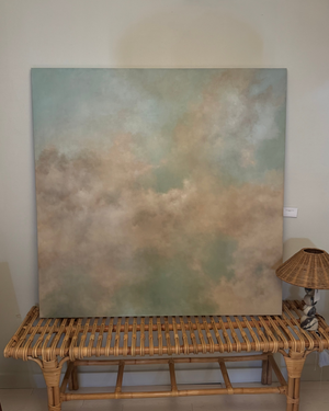 A large square abstract canvas painting with soft blended tones of sage green, beige, and cream, displayed leaning against a wall on a woven rattan bench with a small lamp beside it.