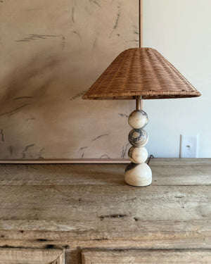 A rustic table lamp with a woven wicker shade and a stacked base made of cream, gray, and brown wooden beads, set on a weathered wooden cabinet.