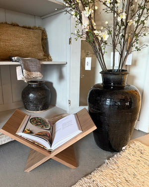A large black glazed ceramic vase filled with flowering branches stands on the floor in a styled interior. A smaller dark ceramic pot sits on a shelf above, with a wooden book stand holding an open book in the foreground.