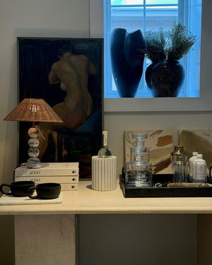 A styled interior scene with a console table holding a woven lamp shade, stacked books, black cups, a ribbed white bottle holder, glassware, and a dark tray. A large framed painting of a nude figure stands against a wall beside a window with blue-tinted light and dark vases.