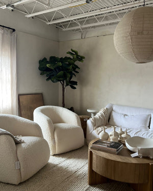 A styled living room featuring two cream boucle armchairs, a white sofa, a round wooden coffee table with ceramic vessels, a large white paper lantern pendant light, and a tall fiddle-leaf fig plant against a warm beige plaster wall.