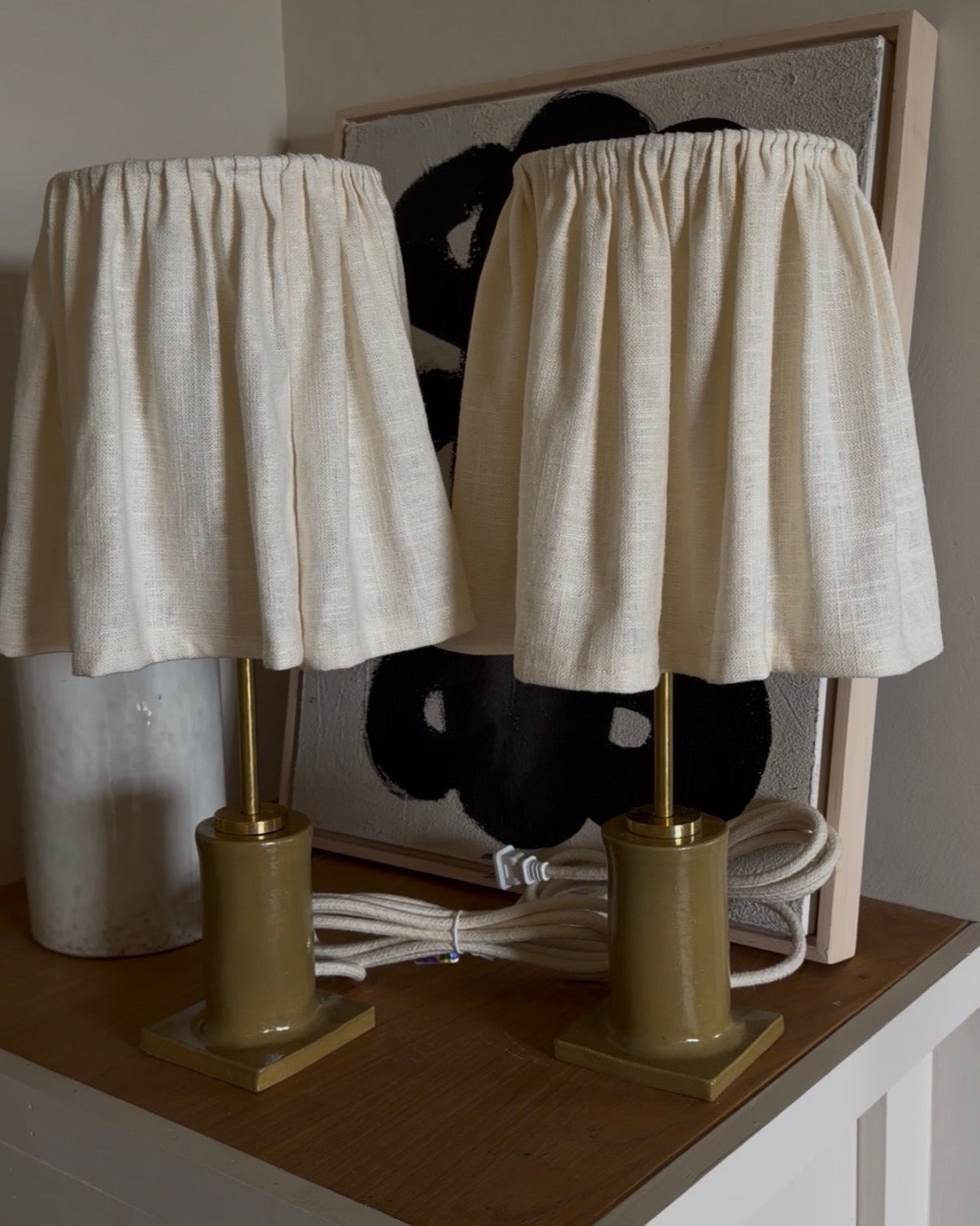 Two table lamps with cream linen shades and gold bases, displayed on a wooden side table in front of a framed abstract print.
