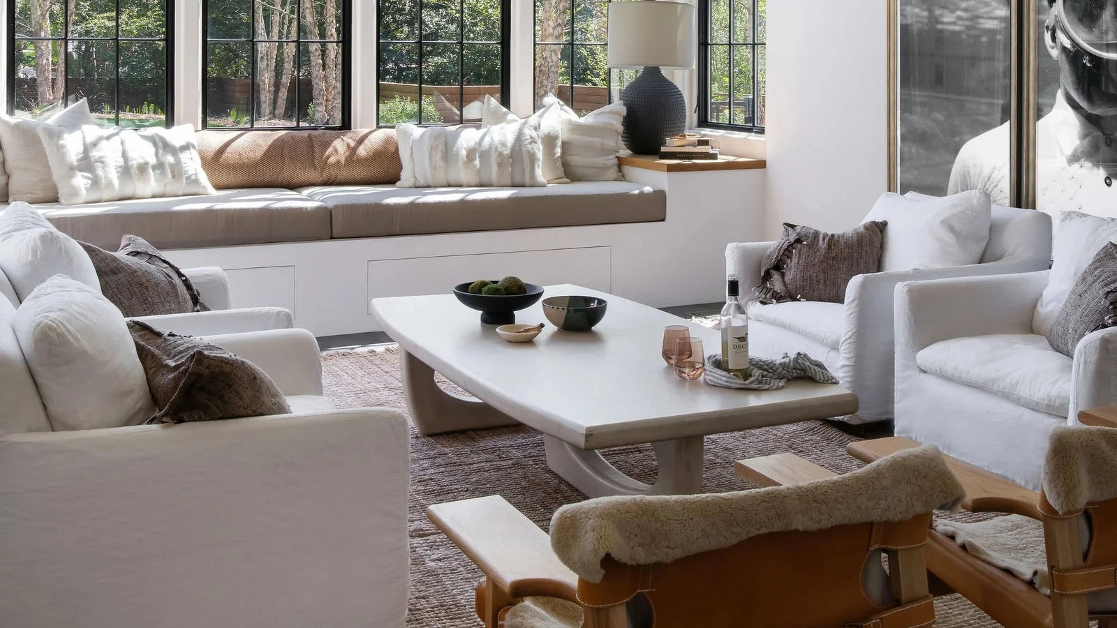 A bright, modern living room with large white cylindrical pendant lights, white sofas, a light wood coffee table, and a woven rug, with large windows showing a view of green trees.