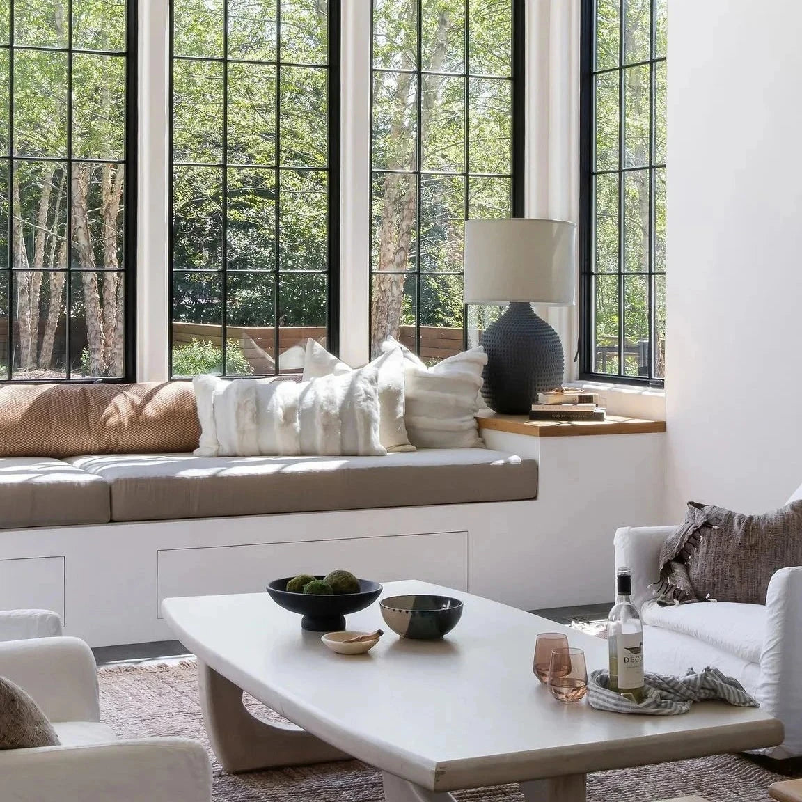 A bright, modern living room with large white cylindrical pendant lights, white sofas, a light wood coffee table, and a woven rug, with large windows showing a view of green trees.